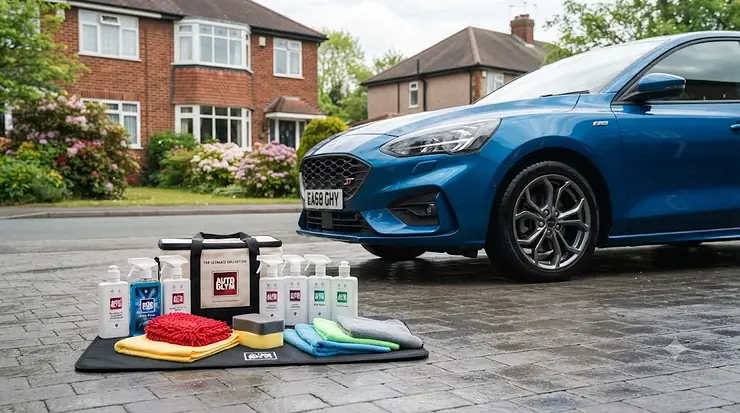 A complete car cleaning kit laid out on a driveway next to a hatchback, featuring shampoo, microfibre cloths, and wheel cleaner. car cleaning kit