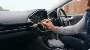 Interior dashboard cleaner and soft-bristle brush removing dust from air vents on a UK right-hand drive car.