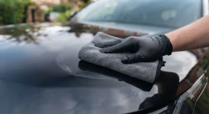 Close-up of a hand in a black nitrile glove using a plush grey microfiche cloth to gently buff away hazy paste wax residue from the black car bodywork, revealing a deep, mirror-like high-gloss finish.