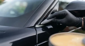 Macro shot of a hand in a black nitrile glove using a small, precise grey foam detailing swab to apply paste wax to the intricate black trim and rubber seal near the A-pillar of the Range Rover Velar.