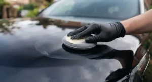 Close-up of a hand in a black nitrile glove using a grey foam applicator pad to apply a thin layer of creamy carnauba paste wax in circular motions to the highly polished black bonnet of the Range Rover Velar.